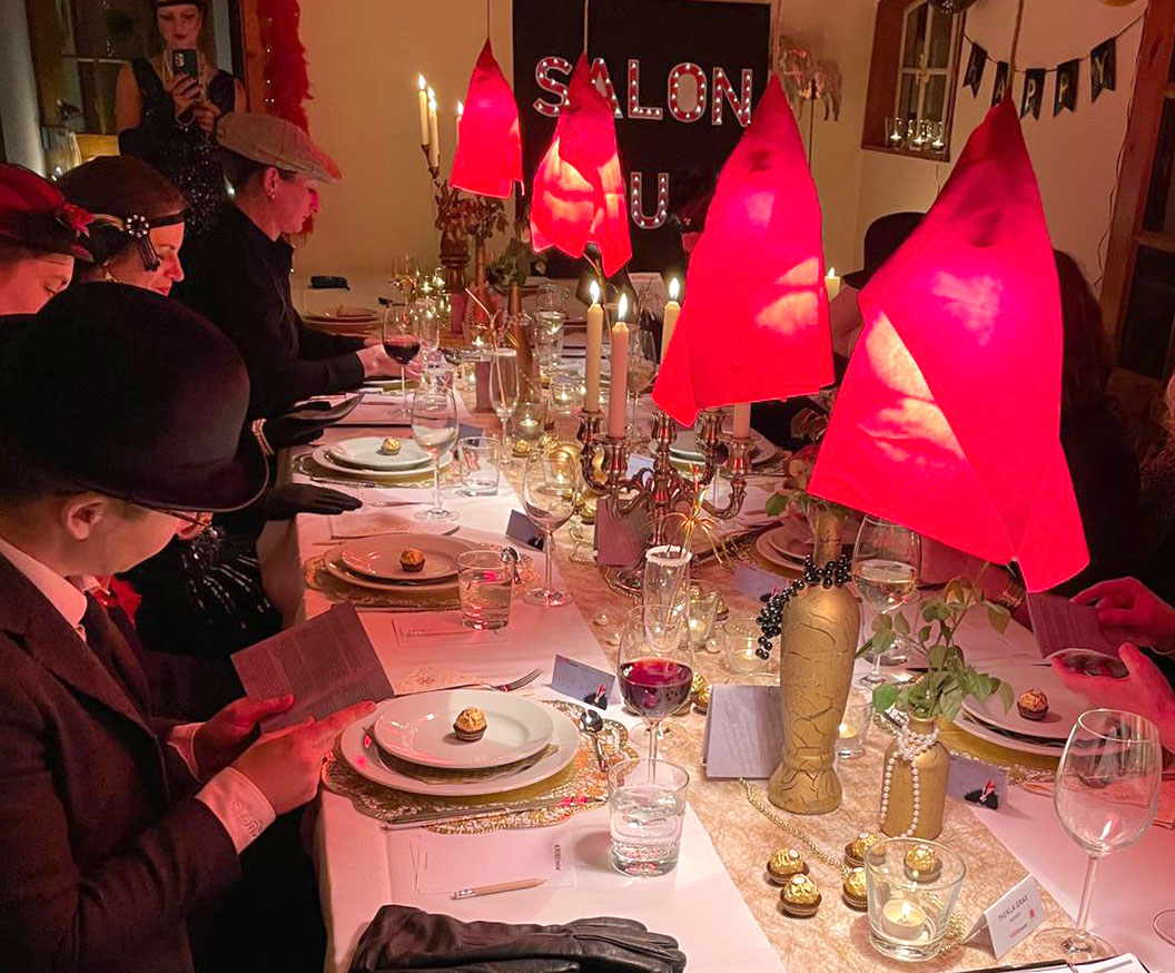 Kostümierte Menschen, die an einem schön dekorierten Tisch sitzen und "Nachts im Salon Rouge" spielen