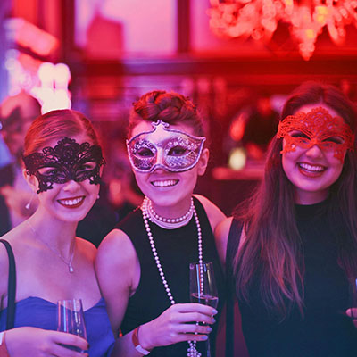 Foto von drei lächelnden Frauen in einer Eventlocation, die Maske tragen und Sektgläser in der Hand halten