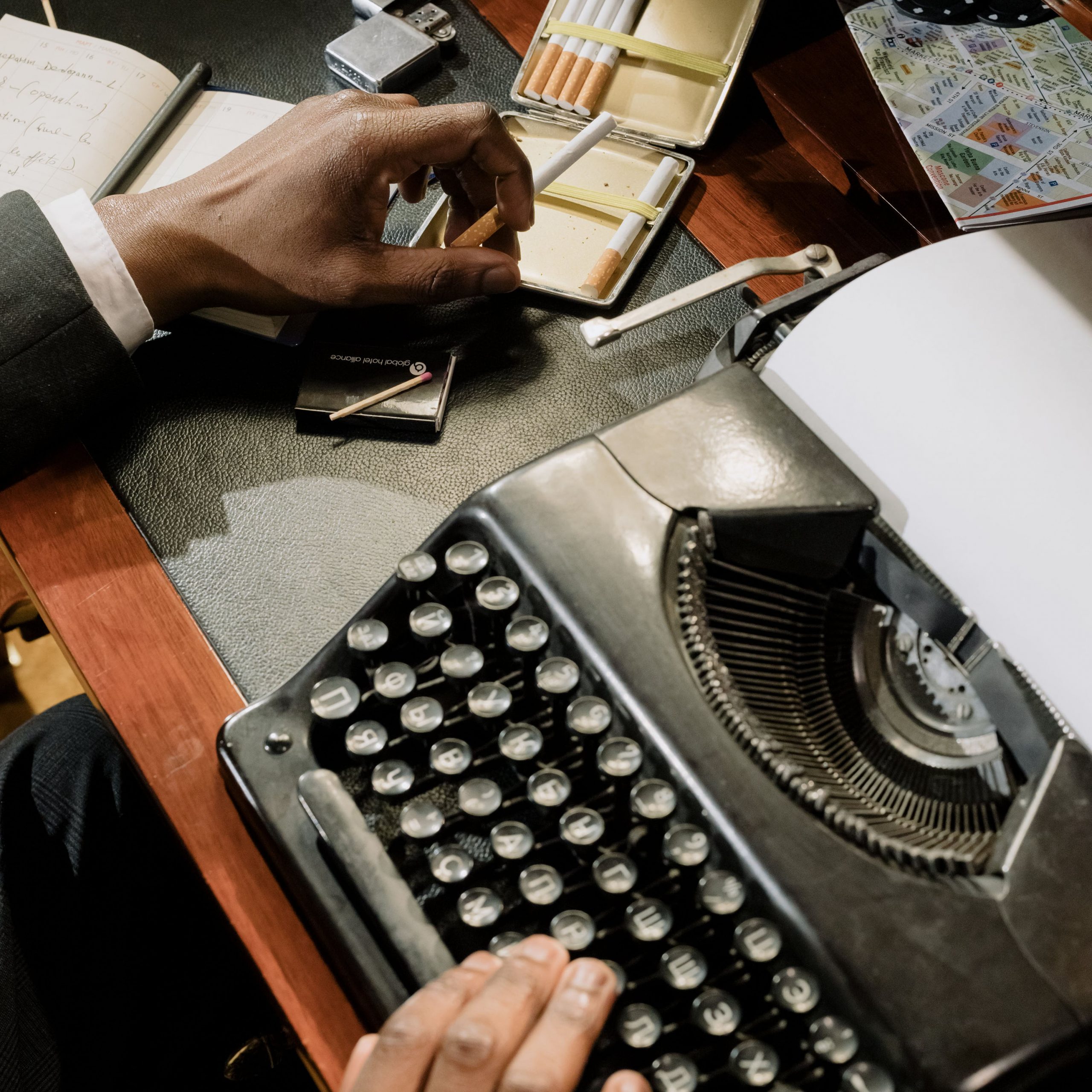 Closeup einer alten Schreibmaschine und Anschnitt einer Hand mit Zigarette sowie Notizbuch und Lageplan