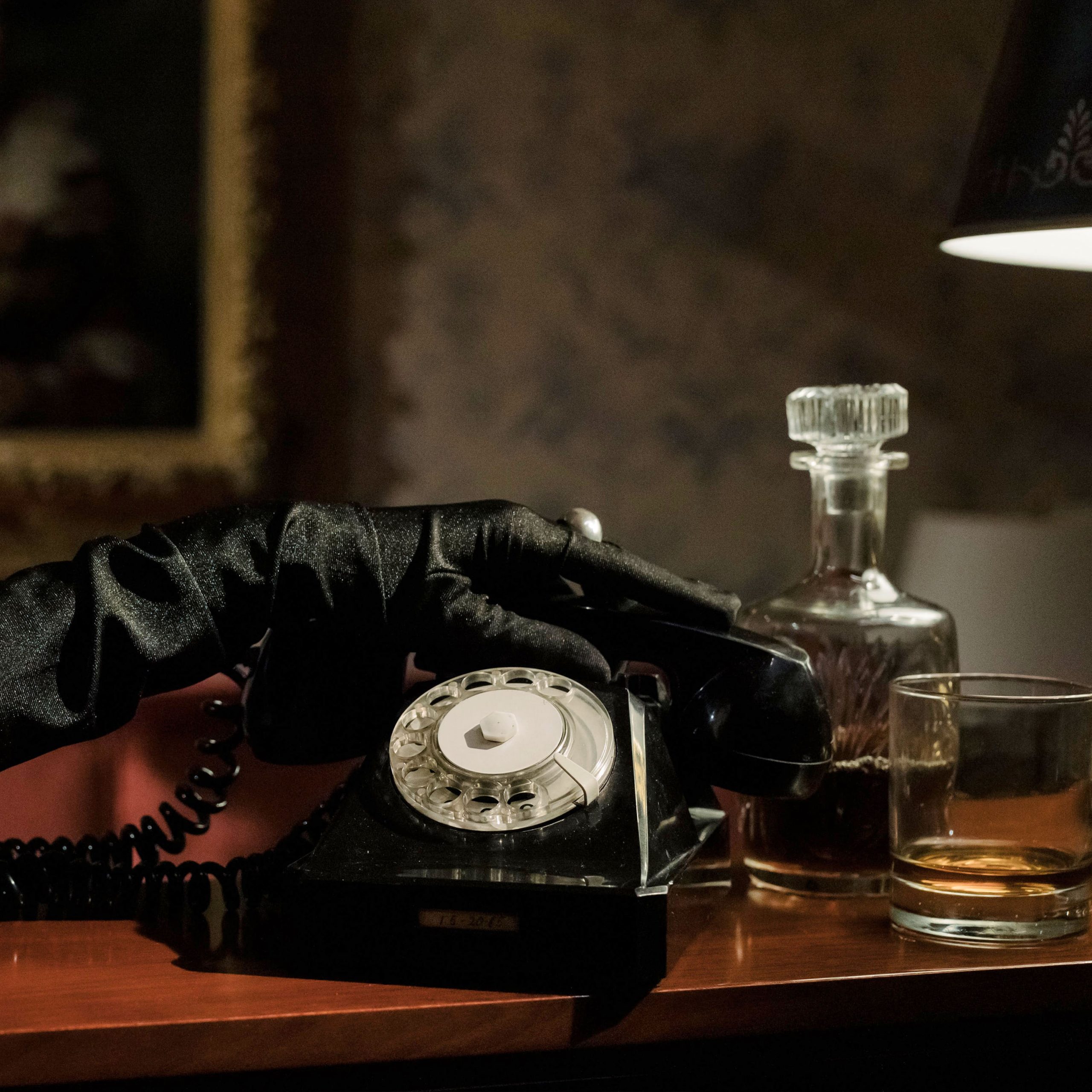 Closeup eines altmodischen Telefons und eine Hand, die zum Hörer greift, welche schwarze Seidenhandschuhe trägt. Daneben ein Whiskeyglas und -flasche.
